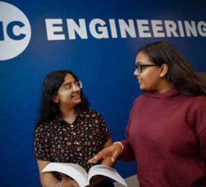two female students talk in the Engineering Innovation Building hallway, against a blue wall with the UIC Engineering logo