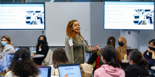 a young mentor in the computing field speaks to female and nonbinary students with two large panel monitor displays in the background