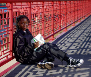 Tomi Babalola sits with a notebook and pen on a red metal bridge that crosses the Chicago river