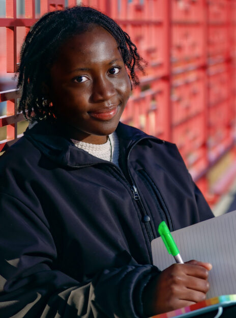 Tomi Babalola sits with a notebook and pen on a red metal bridge that crosses the Chicago river