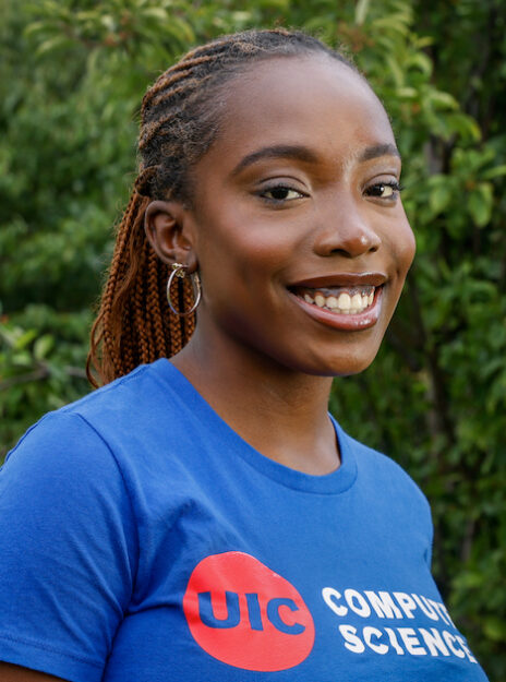 Tomi Okunola poses in a royal blue UIC computer science t-shirt against a background of green leaves