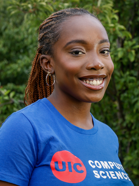 Tomi Okunola poses in a royal blue UIC computer science t-shirt against a background of green leaves